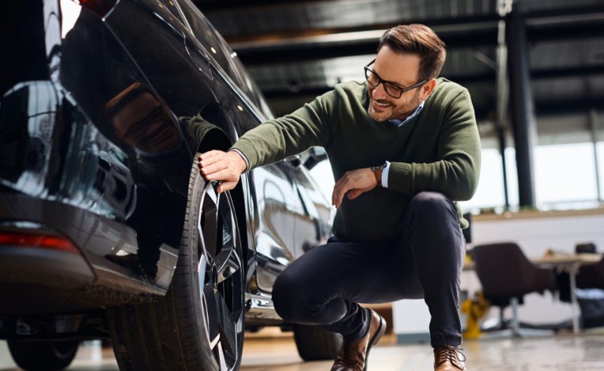 A happy man running his hand over the tire on a vehicle at a car dealership. He is kneeling next to the car.