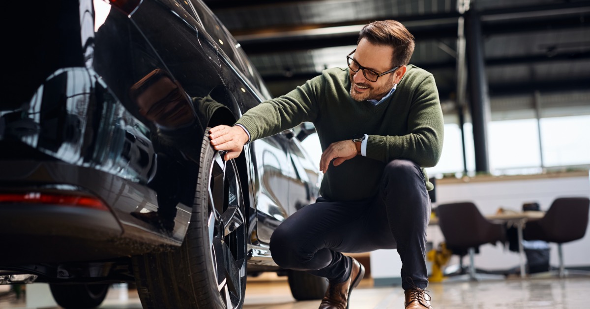 A happy man running his hand over the tire on a vehicle at a car dealership. He is kneeling next to the car.
