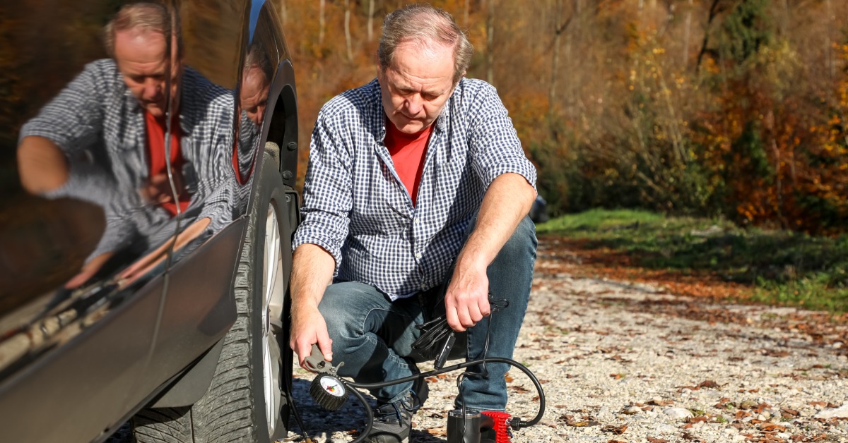 A man in a checkered shirt and jeans is looking down at his portable tire pump, which is attached to his tire.