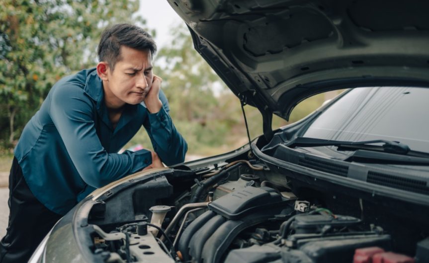 A man leaning his elbows and propping his head up while examining the open engine bay of his car during the day.