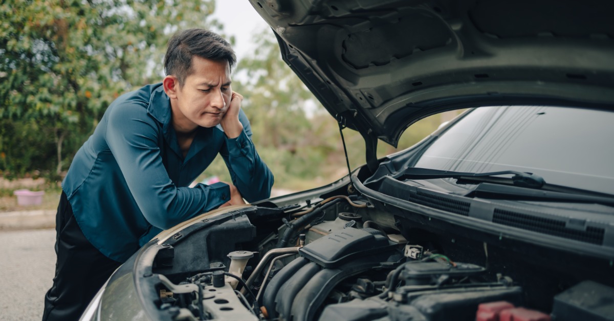 A man leaning his elbows and propping his head up while examining the open engine bay of his car during the day.