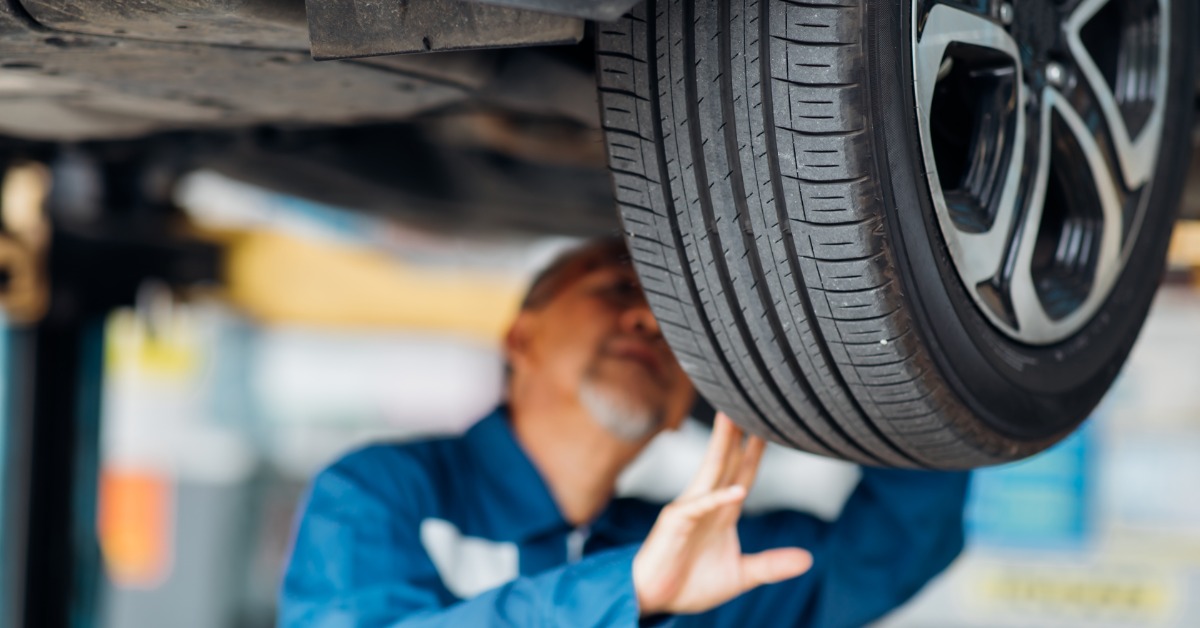 A close-up of the rear tire of a car elevated on a lift with a blurred man in a blue jumpsuit examining it.