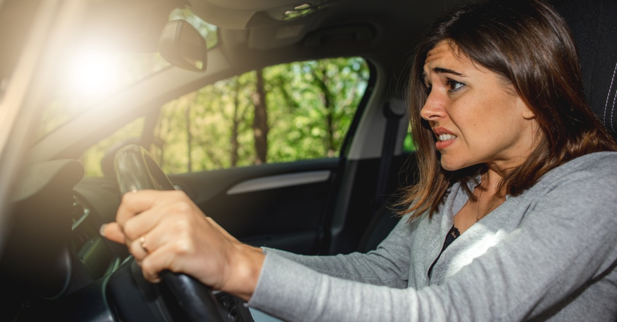 A side view of a female driver in a grey sweater, grimacing and gripping the steering wheel of her car tightly.
