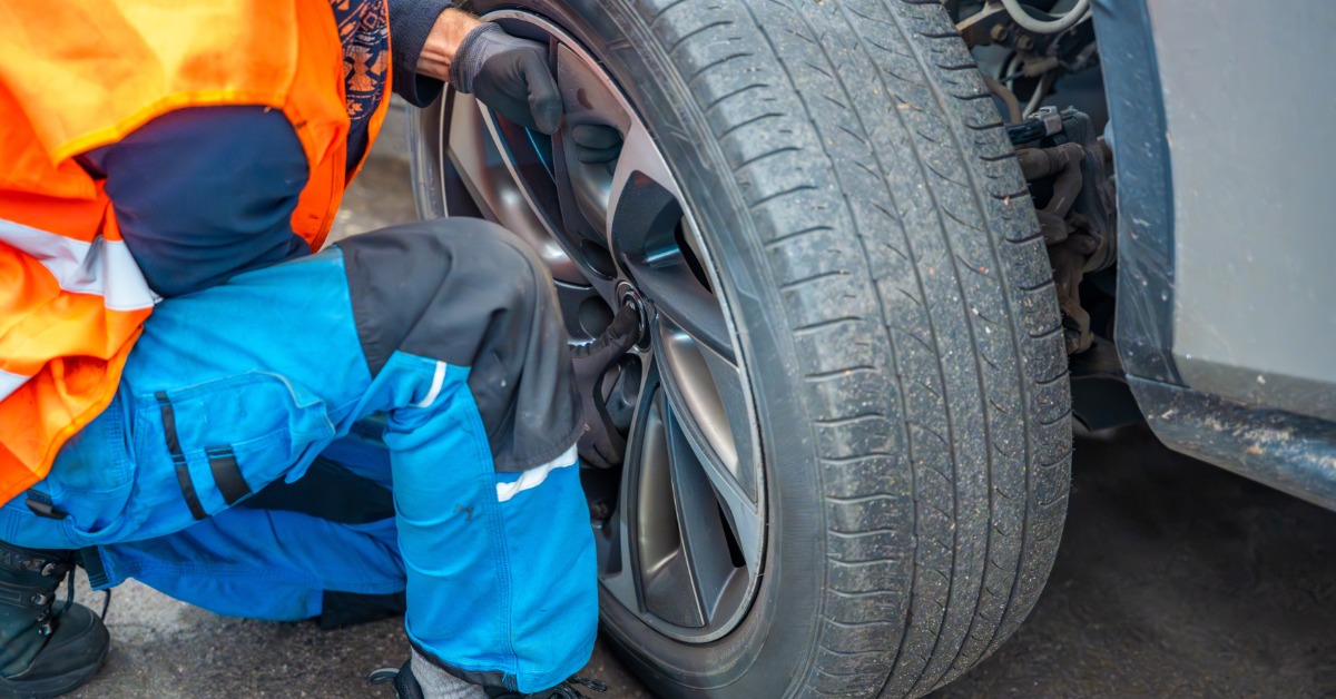 Someone wearing an orange safety vest kneels next to a vehicle. They lift a tire onto one of the car's axles.