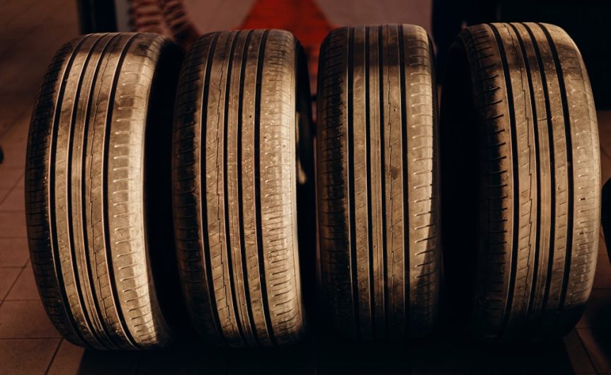 A set of four used tires propped up on a shop floor with the sun casting over them. Each tire is worn differently.