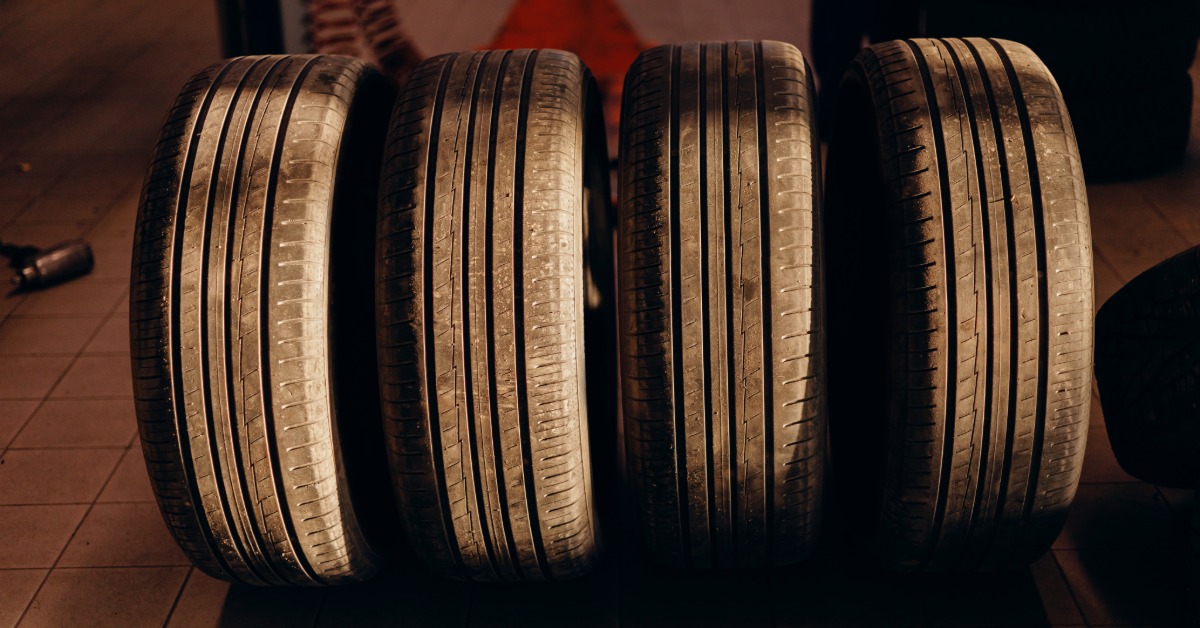 A set of four used tires propped up on a shop floor with the sun casting over them. Each tire is worn differently.