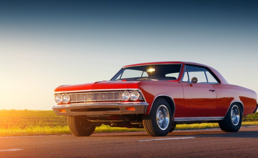 A red classic car on a two-lane highway with grass in the background and the sun setting out of frame.
