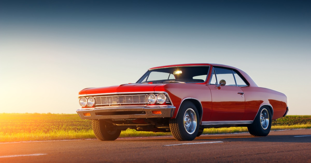 A red classic car on a two-lane highway with grass in the background and the sun setting out of frame.