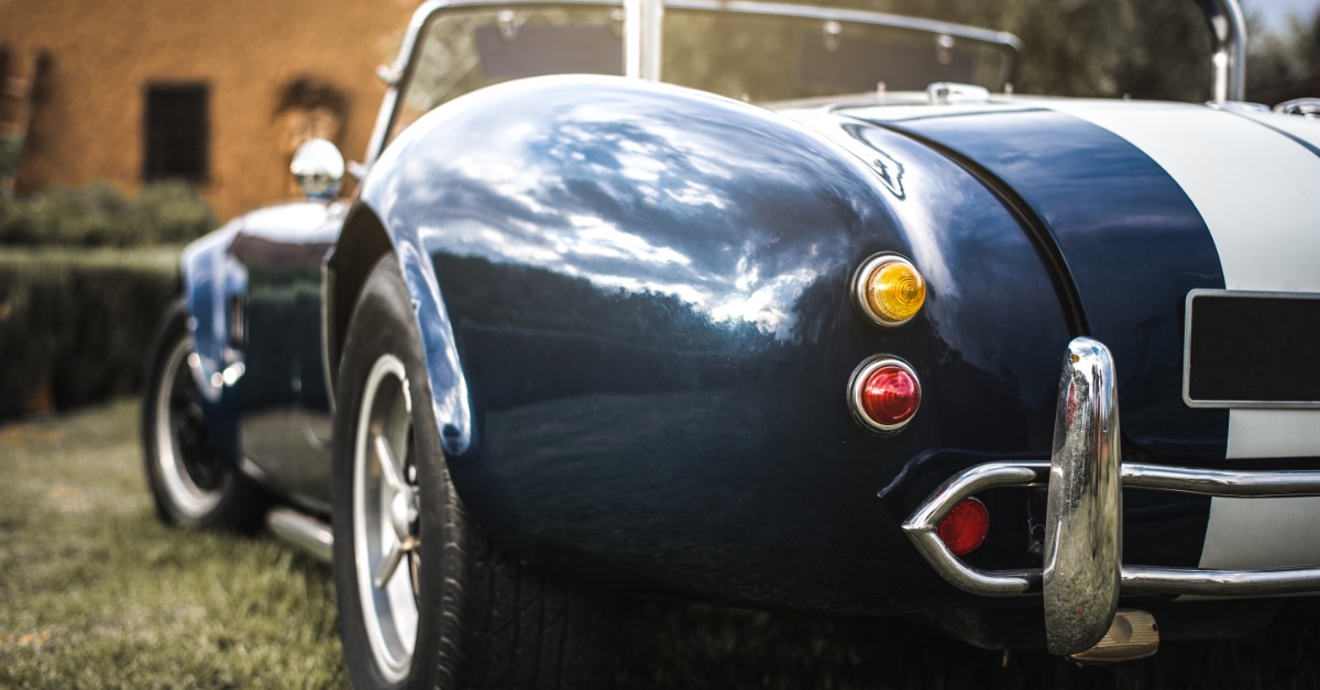 A rear view of a blue classic soft top sports car with the top down and a white racing stripe parked on grass.