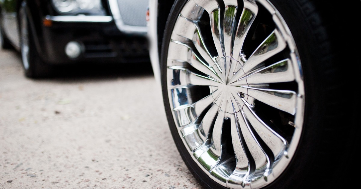 A close-up of a white car's shiny chrome wheel parked on a pavement with a black car opposite facing it.
