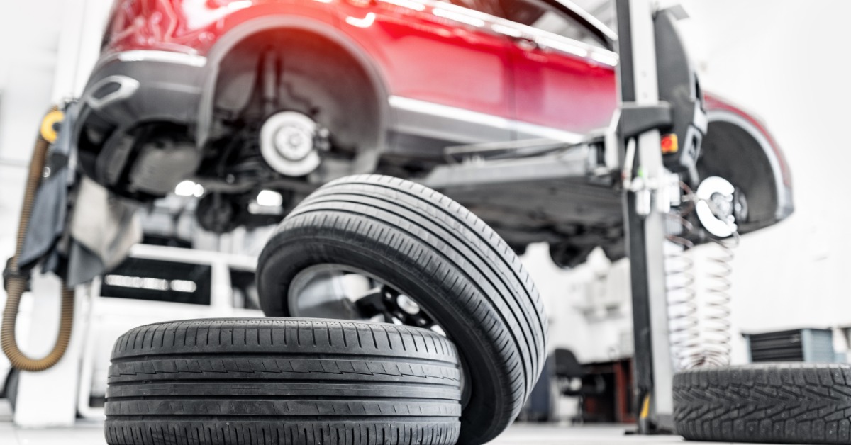 A couple of tires lie haphazardly on the ground of a shop floor. A red SUV is lifted with no tires on.