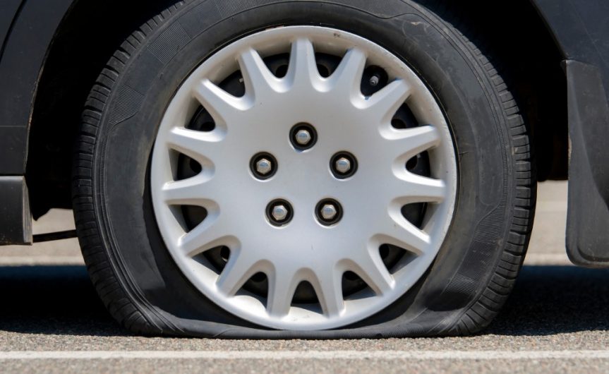 A close-up side view of a wheel with a flat tire attached to a black car resting on pavement with white lines.
