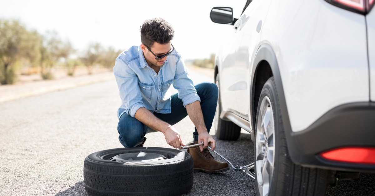 A man in a denim shirt and jeans with sunglasses using a jack to raise a white car with a spare tire next to him.