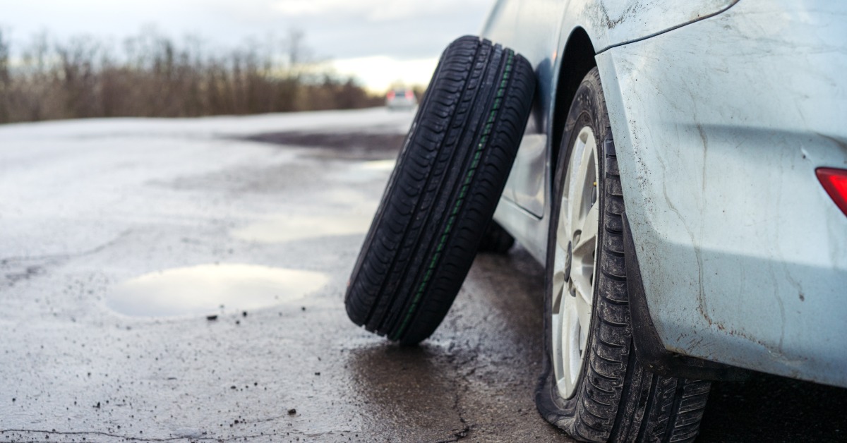 A close-up of the rear end of a grey tire with a flat rear tire, with a new spare tire leaning against it.