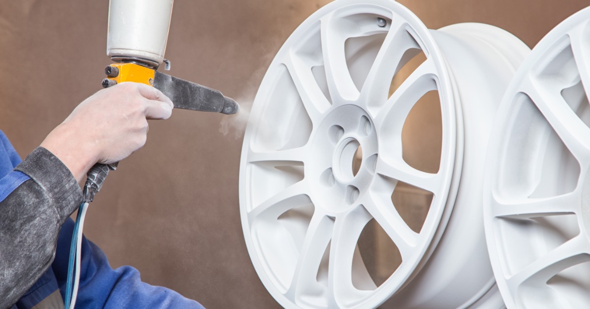 Someone wearing gloves holds a car wheel in one hand and sprays it with a powder coating. Another wheel rests to the right.