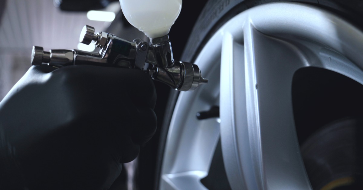 A close-up of someone holding a spray gun filled with paint up to the wheel of a car. The person wears black latex gloves.