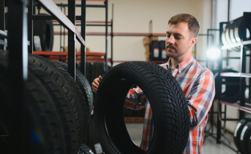 A man wearing a blue and red flannel stands in a tire shop. He lifts a tire from a rack and inspects it.