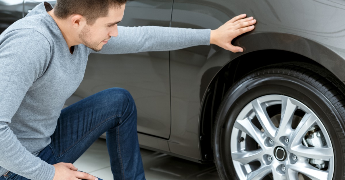 A man kneels next to the side of his car by the tires. One hand rests on the vehicle while he looks at the tire.