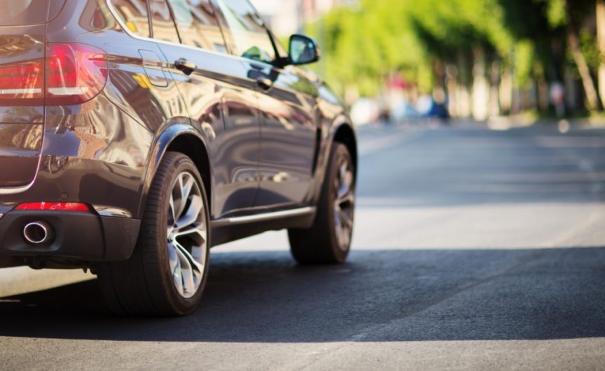 A rear view of a dark midsize crossover SUV driving on a city street on a sunny day with trees in the background.