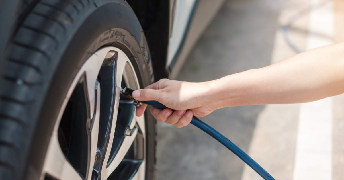 A close-up of a woman's hand holding a blue air pressure hose and nozzle to the stem of a car tire to inflate it.