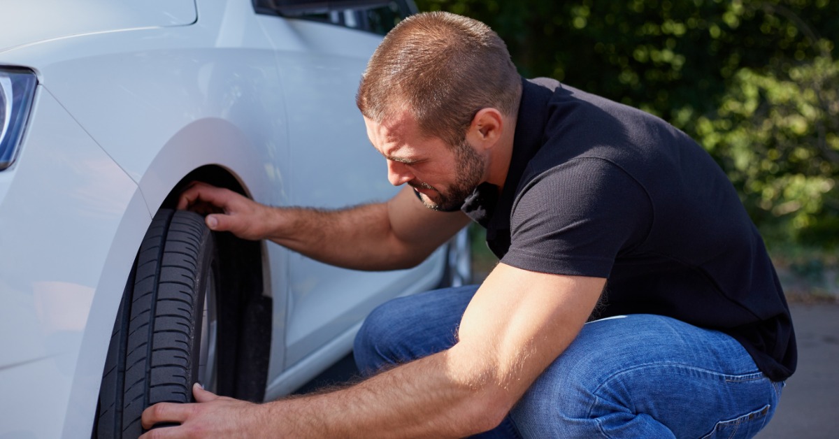 A man in a black t-shirt and blue jeans crouches down to examine the front tires of a white car during the day.