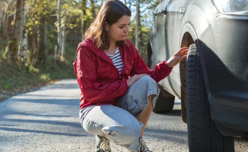 A woman kneels next to the front tire of her car. One hand touches the tread as she looks at the tire.