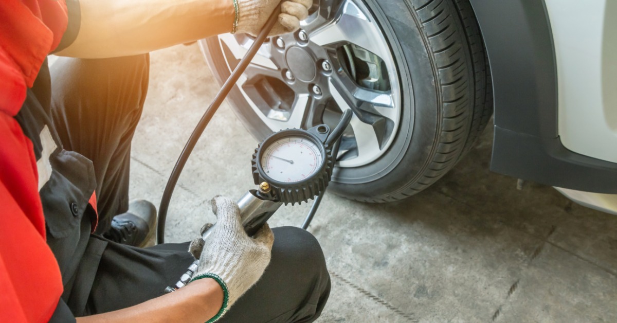 A man in a red shirt and black pants with white gloves crouches next to a tire to use an air pressure gauge.