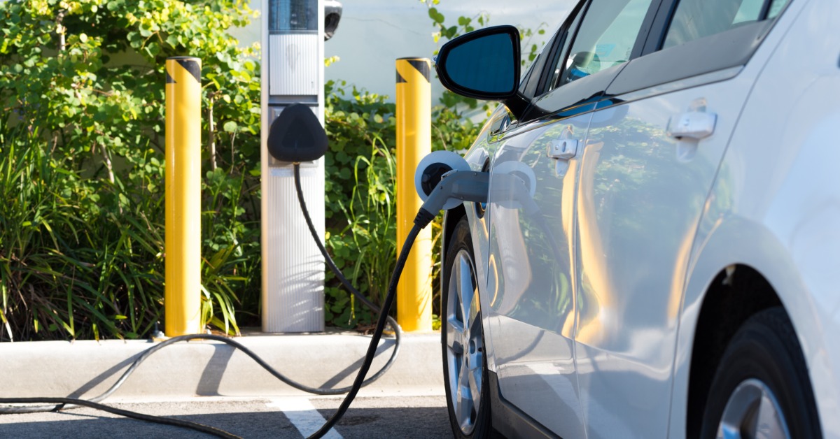 A close-up of the side of a white electric vehicle parked in a parking lot and connected to a public charging station.