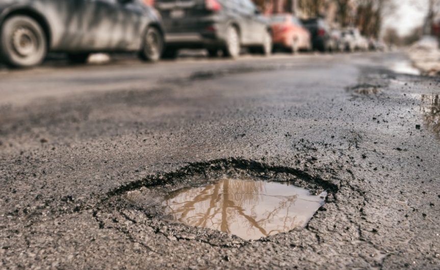 A close-up of a pothole filled with muddy water. Cars are blurred and parked along the street behind the pothole.
