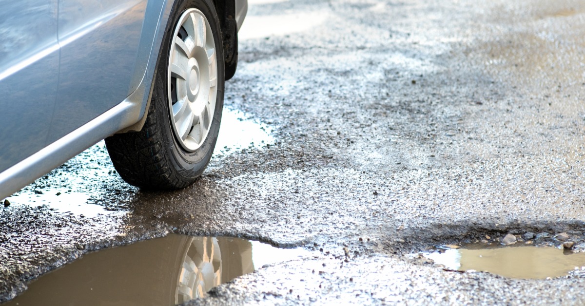 Two large potholes next to each other and filled with muddy water. A blue car's back tire rolls next to one of the potholes.