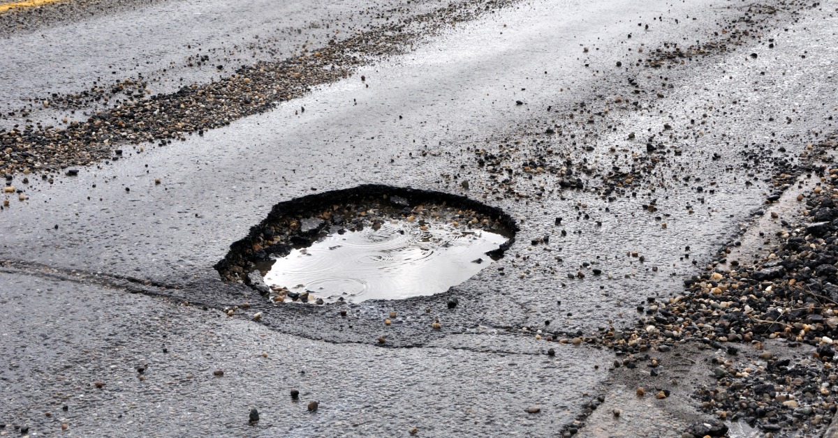 A very deep pothole in the middle of a road. Broken asphalt and gravel are scattered across the road, too.