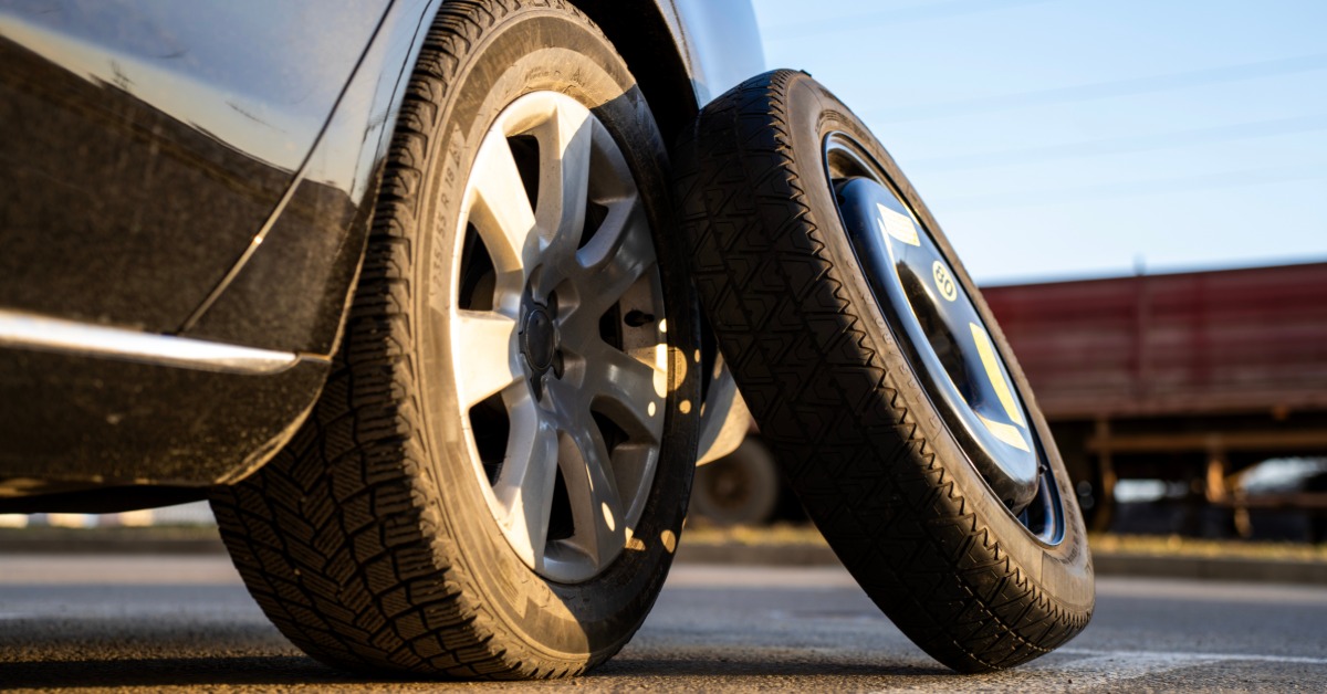 A close-up of the rear wheel and tire of a car with a spare wheel propped up against it outside in a parking lot.