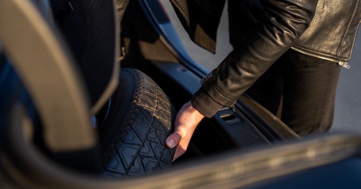A close-up of a man in a leather jacket as he opens the luggage compartment of a car's trunk to remove the spare tire.