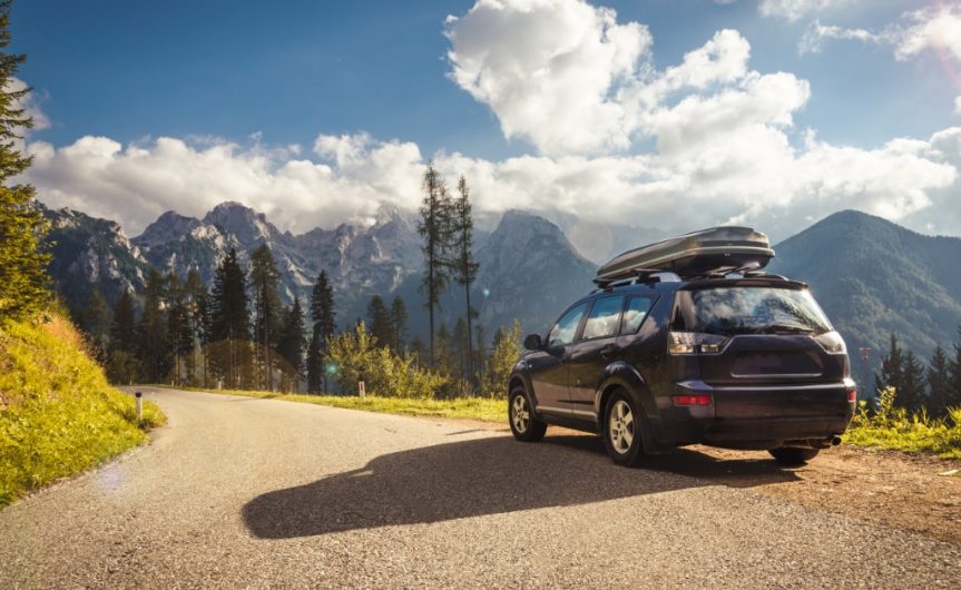 A black SUV with a roof rack parked on the dirt shoulder of a road. Mountains and clouds fill the sky.