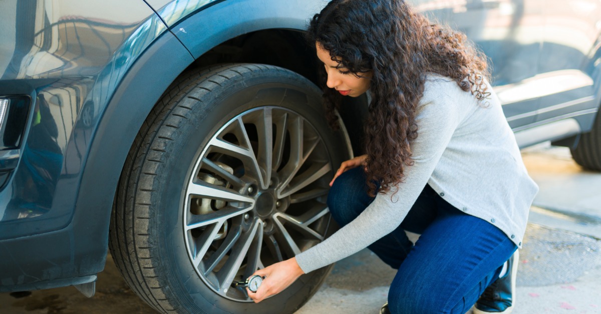 A woman kneels next to the front tire of her vehicle. She uses a pressure gauge to check the PSI of the tire.