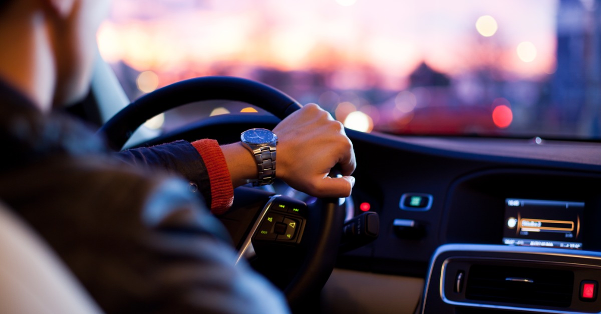 A close-up of someone sitting behind the wheel of a vehicle. Their left arm rests on the steering wheel.