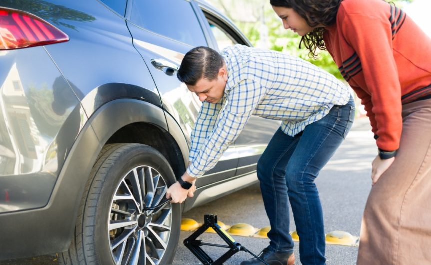 A man crouching down with a tool to loosen the lug nuts of a car's tire outside while a young girl watches.
