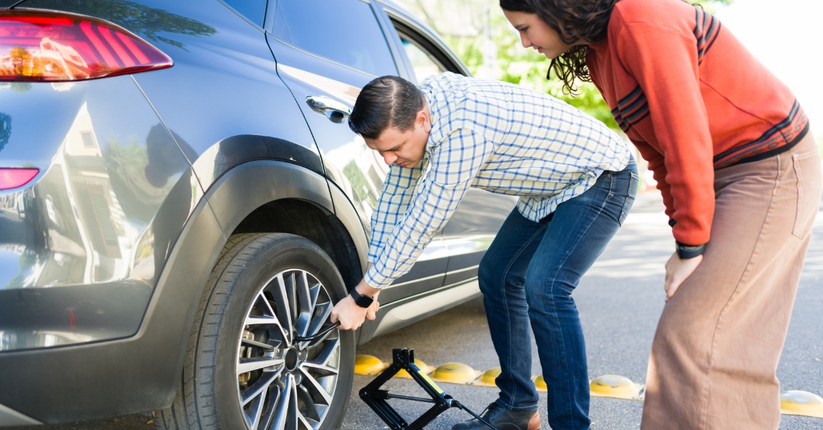 A man crouching down with a tool to loosen the lug nuts of a car's tire outside while a young girl watches.