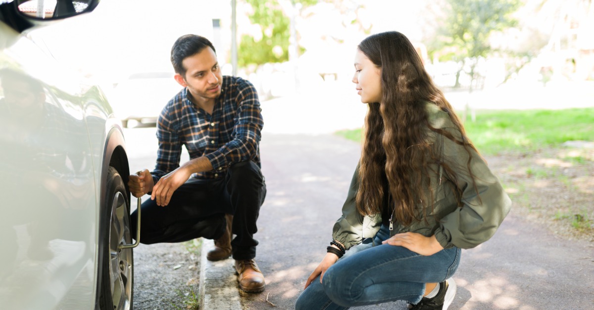 A man crouches on a curb while using a wrench to loosen the lug nuts of a tire and talks to a young girl.