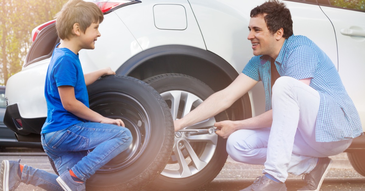 A man and a young boy smile as the man changes the tire of a white SUV, and the boy holds the spare tire.