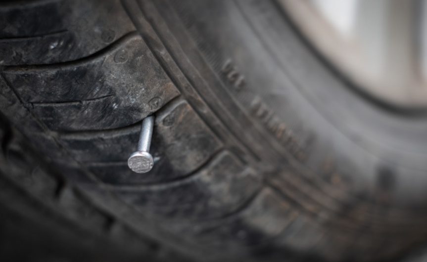 A close-up of the head of a nail sticking out of the tread of an automotive tire, with the rest of the frame blurred.