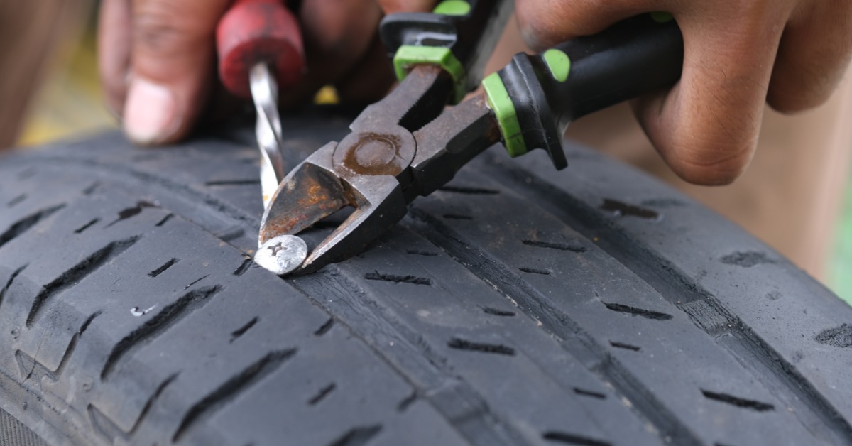 A close-up of a pair of hands using a screwdriver and a pair of pliers to remove a nail in a car tire.