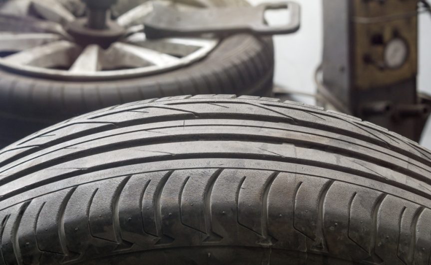 A close-up of the tread of an individual, upright car tire. The used tire shows signs of cracking and dry rot.
