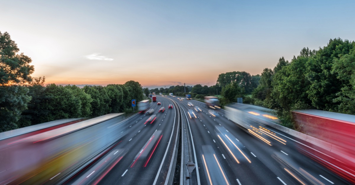 A blur of vehicles driving at a fast speed on a highway. The sky above the highway is clear with no clouds.