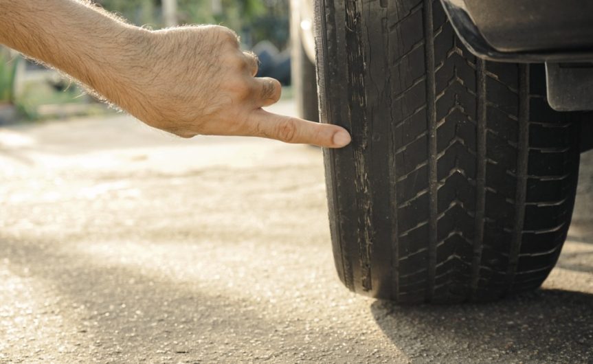 A man's hand pointing at the damage on a worn-out tire. The tread is worn down and balding in various spots.