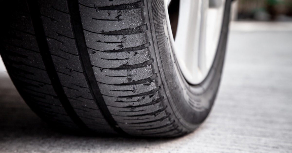 A tire attached to an old car with damage along various parts of the tread. The tread is balding and partially cut.