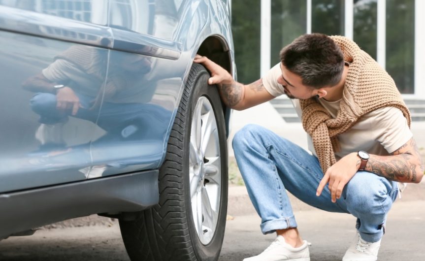 A well-dressed man crouched next to a pale blue car. He has one hand on the tire while looking at it.