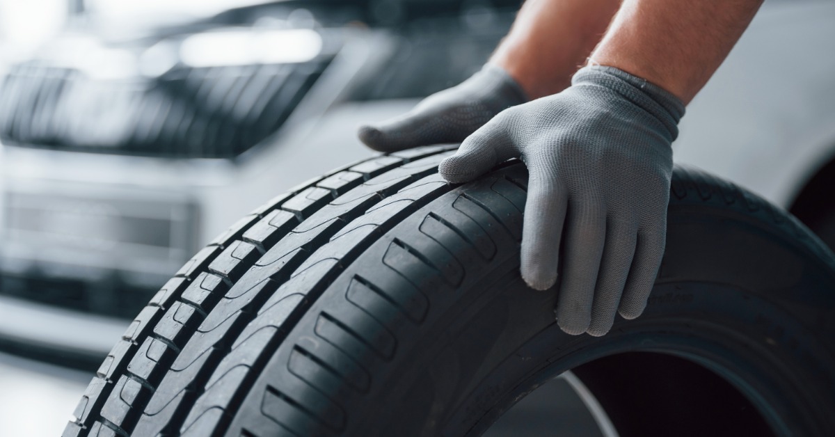 A mechanic in a workshop wearing grey gloves holding a new tire. The tire is clean and has no marks.