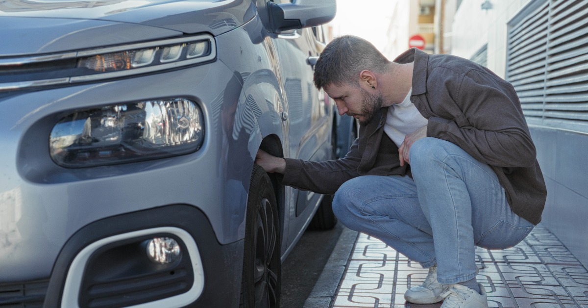 An SUV parked next to a curb. A man kneels next to the front tire, examining it and touching it with his hand.