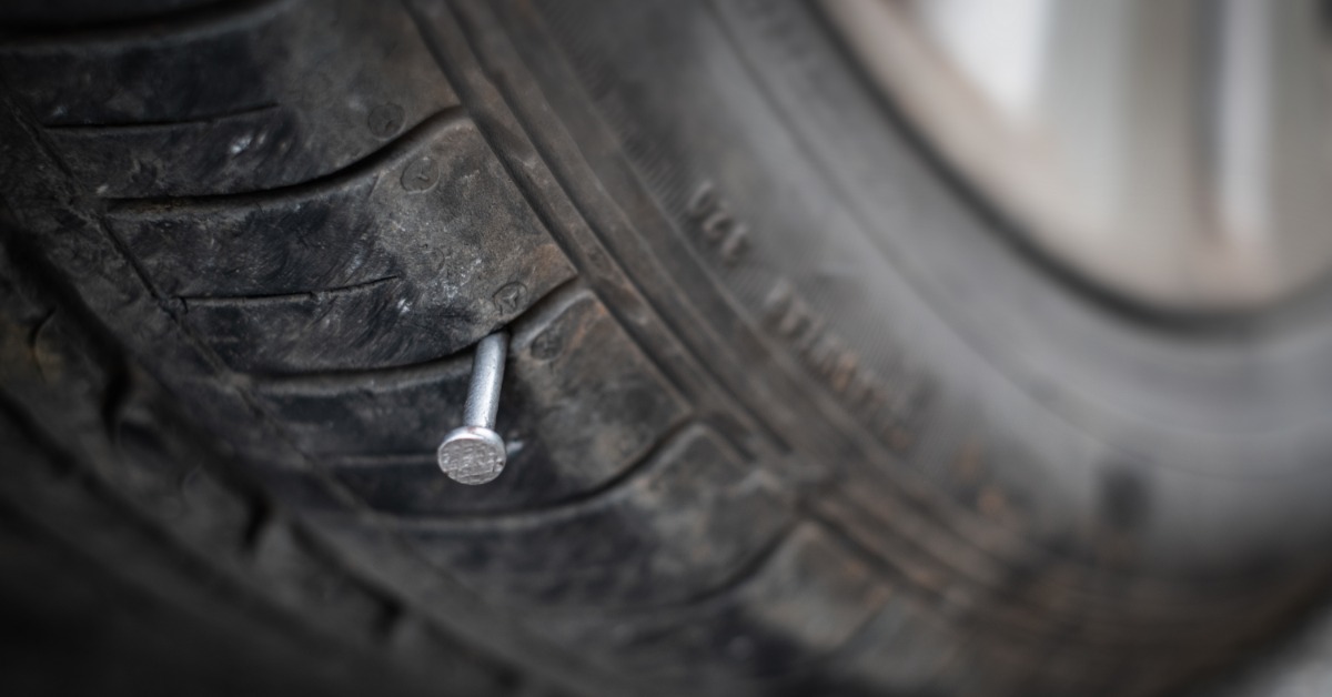 A close-up of the tread of a tire. A small nail punctures the tire and sticks out slightly from the rubber.